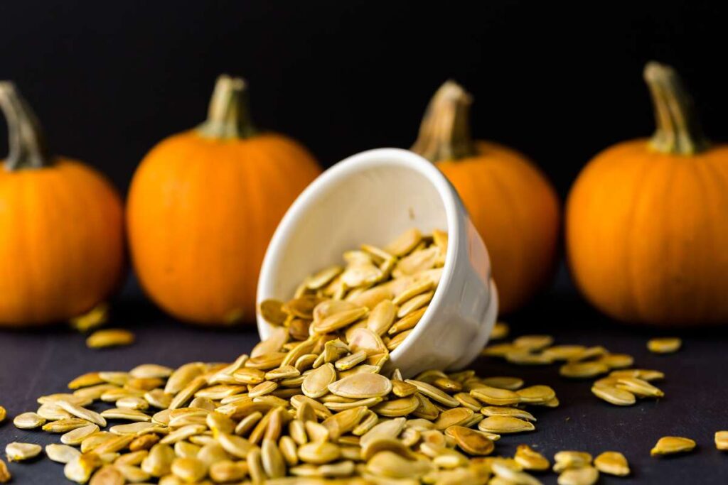 A bowl overflowing with roasted pumpkin seeds, surrounded by fresh pumpkins.