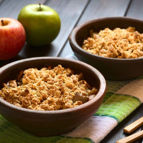 Bowl of warm apple crumble with oats covered in a crisp biscuit topping. The filling is gooey apples with tangy rhubarb.