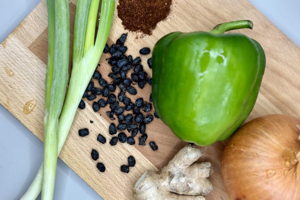 Spring onions, green peppers, onions, ginger, black beans and spices on a chopping board.